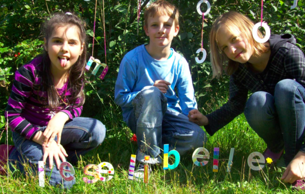 2 Mädchen und 1 Junge sitzen auf grüner Wiese, vor den 3 Kindern steckt ein bunter Schriftzug "Leselibelle" in der Wiese.