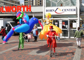 3 Walking-Acts (ein Vogel, ein Clown auf dem Fahrrad und eine Frau in gelbem Kleid) stehen vor dem Haupteingang des Born Centers