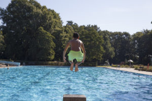 ein Junge mit grüner Badehose springt mit angezogenen Beinen ins Wasser