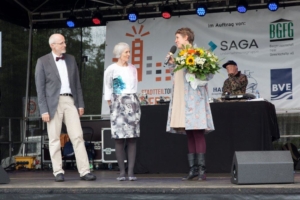 Eine Frau mit Blumenstrauß hält eine Rede, 2 weitere Personen stehen mit auf der Bühne