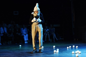 ein verkleideter Mann mit Bommelmütze steht auf einer Bühne. Im dunklen Hintergrund sieht man ein paar Besucher