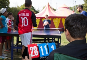 2 Jungen spielen an einer Tischtennisplatte. Im Vordergrund sitzt ein Jungen und zählt die Punkte