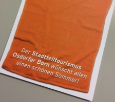 ein orangefarbenes Handtuch liegt auf einem Blatt und dort steht drauf "Der Stadtteiltourismus Osdorfer Born wünscht allen einen schönen Sommer!"