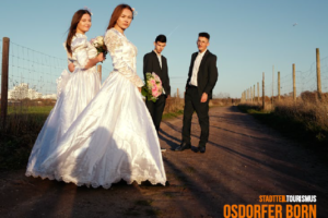 2 Frauen im weißen Hochzeitskleid, die einen bunten Blumenstrauß in der Hand halten und 2 Männer in einem schwarzen Anzug stehen auf einem Feldweg.