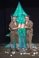 2 Männer in braunen Klamotten tragen ein Rentiergeweih. Sie stehen vor einem grünen Tannenbaum und halten ein Geschenk in der Hand