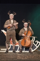 2 Männer in braunen Klamotten tragen ein Rentiergeweih. Der eine sitzt auf einem Schlitten, der andere steht. Beide spielen ein Musikinstrument